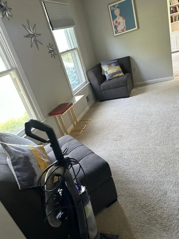 Living room with gray sofa, armchair, large windows, wall art, and a vacuum in the foreground