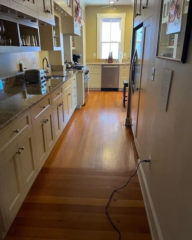 Narrow kitchen with white cabinets, wood floor, and a window at the far end.