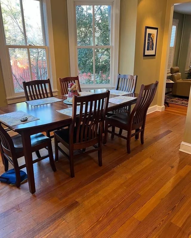 Dining room with wooden table and chairs, hardwood floor, and large windows overlooking trees.