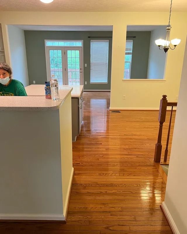 Bright hallway with hardwood floors, white walls, a staircase, and a person partially visible behind a counter.