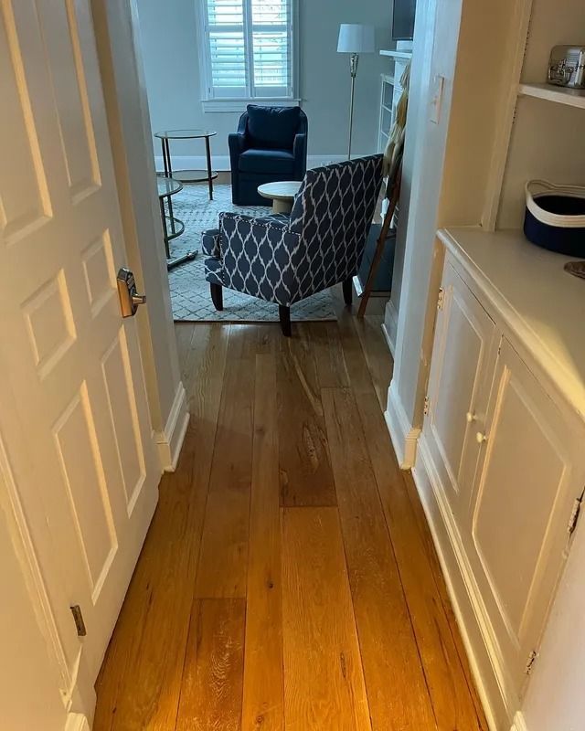 Hallway with wooden floor leading to a living room, featuring a patterned chair and a lamp near a window.