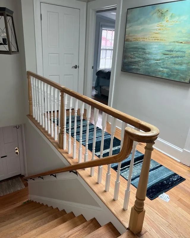 Staircase with wood railing and blue runner rug in a bright hallway, with a large seascape painting on the wall