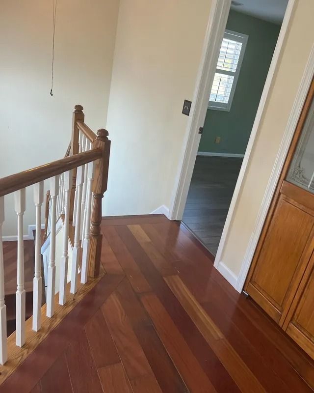 Hallway with wood floors, white walls, staircase railing, and a doorway to a green room.