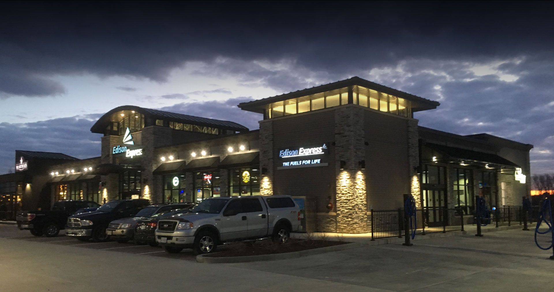 Exterior view of a restaurant complex at dusk. Stone facade with multiple storefronts. Cars are parked in front.