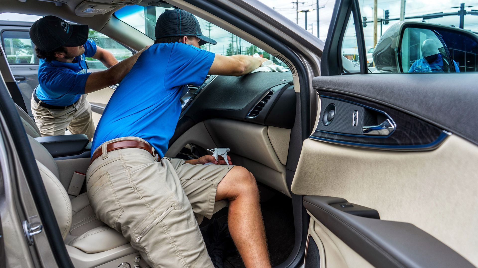 Two people cleaning the interior of a car. One wipes the dashboard, the other cleans something near the door.