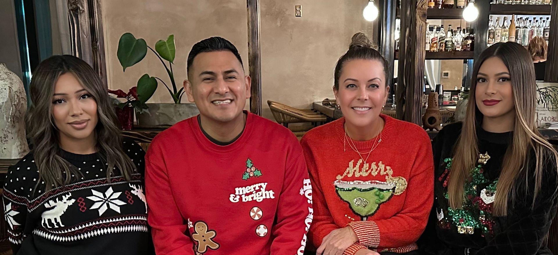 Four people posing indoors, two in red holiday sweaters, with festive decor in the background.