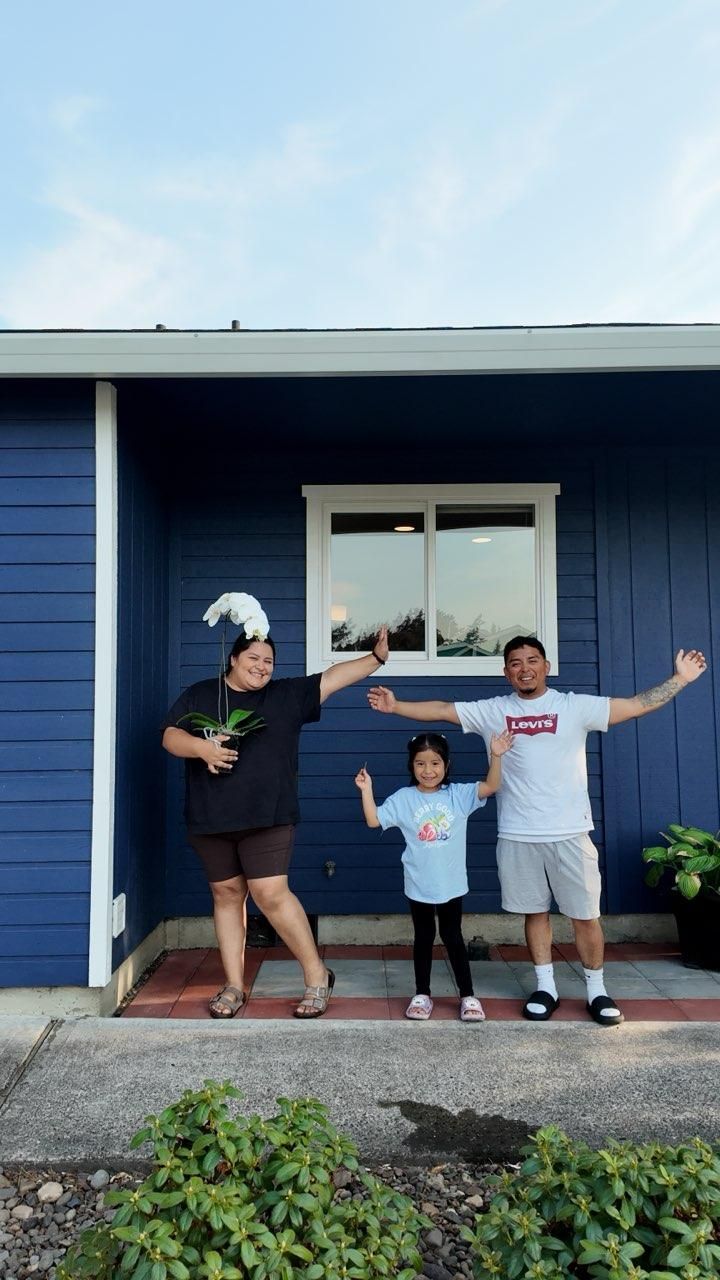 Three people stand smiling outside a blue house with arms outstretched on the porch.