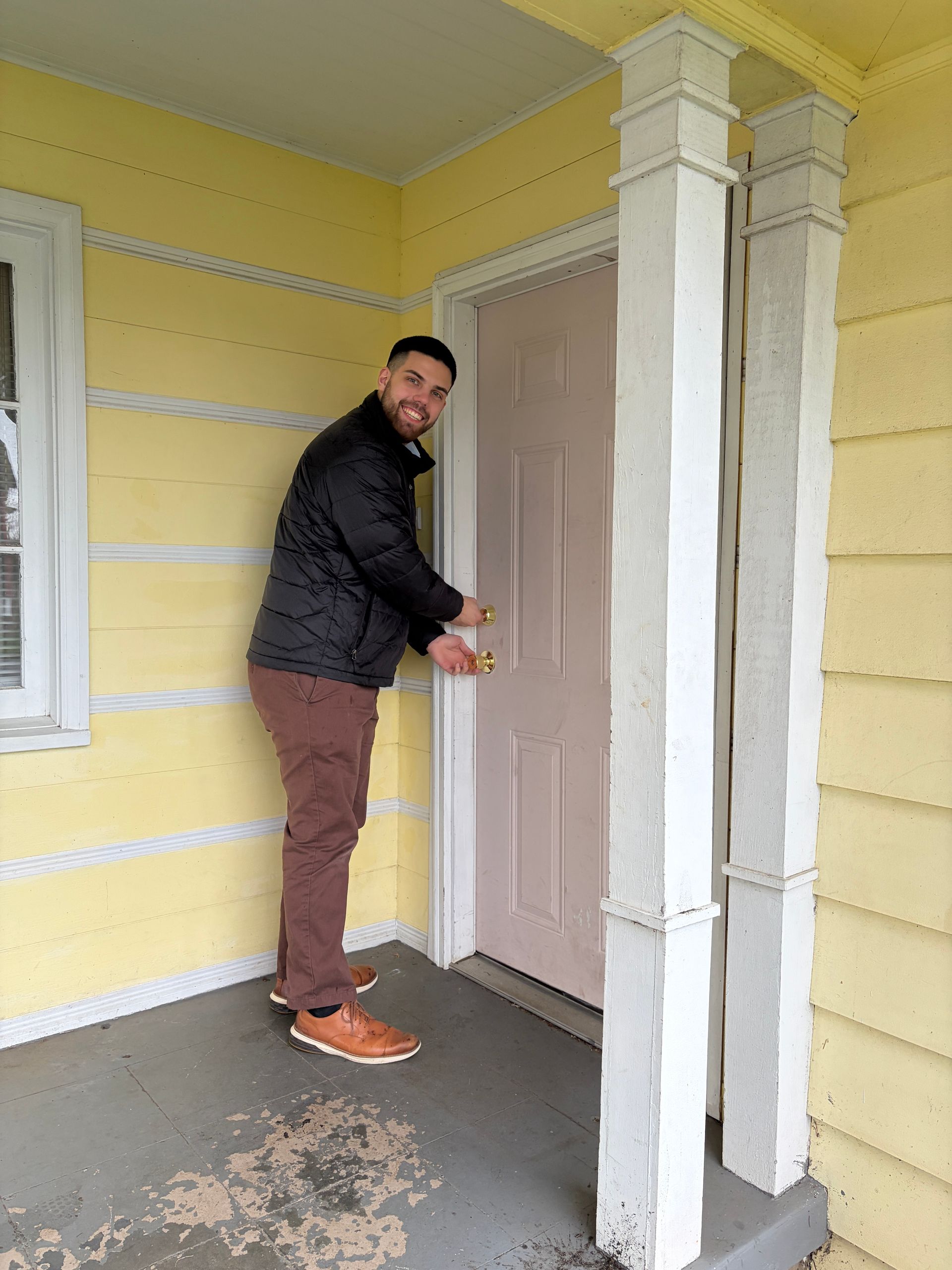 Man in dark jacket unlocking a pink front door on a yellow porch