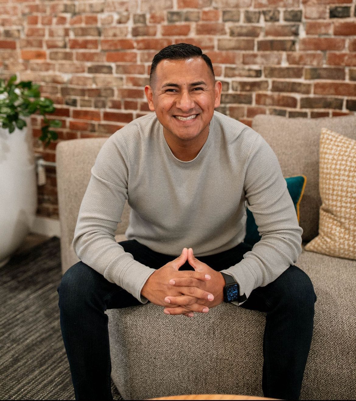 A man is sitting on a couch with his hands folded and smiling.