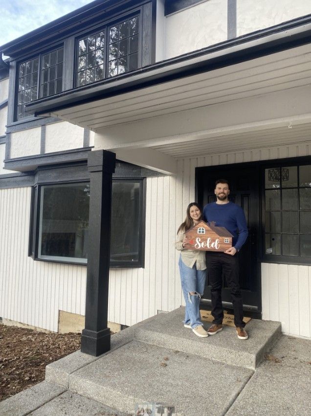 A man and a woman are standing in front of a house holding a sold sign.