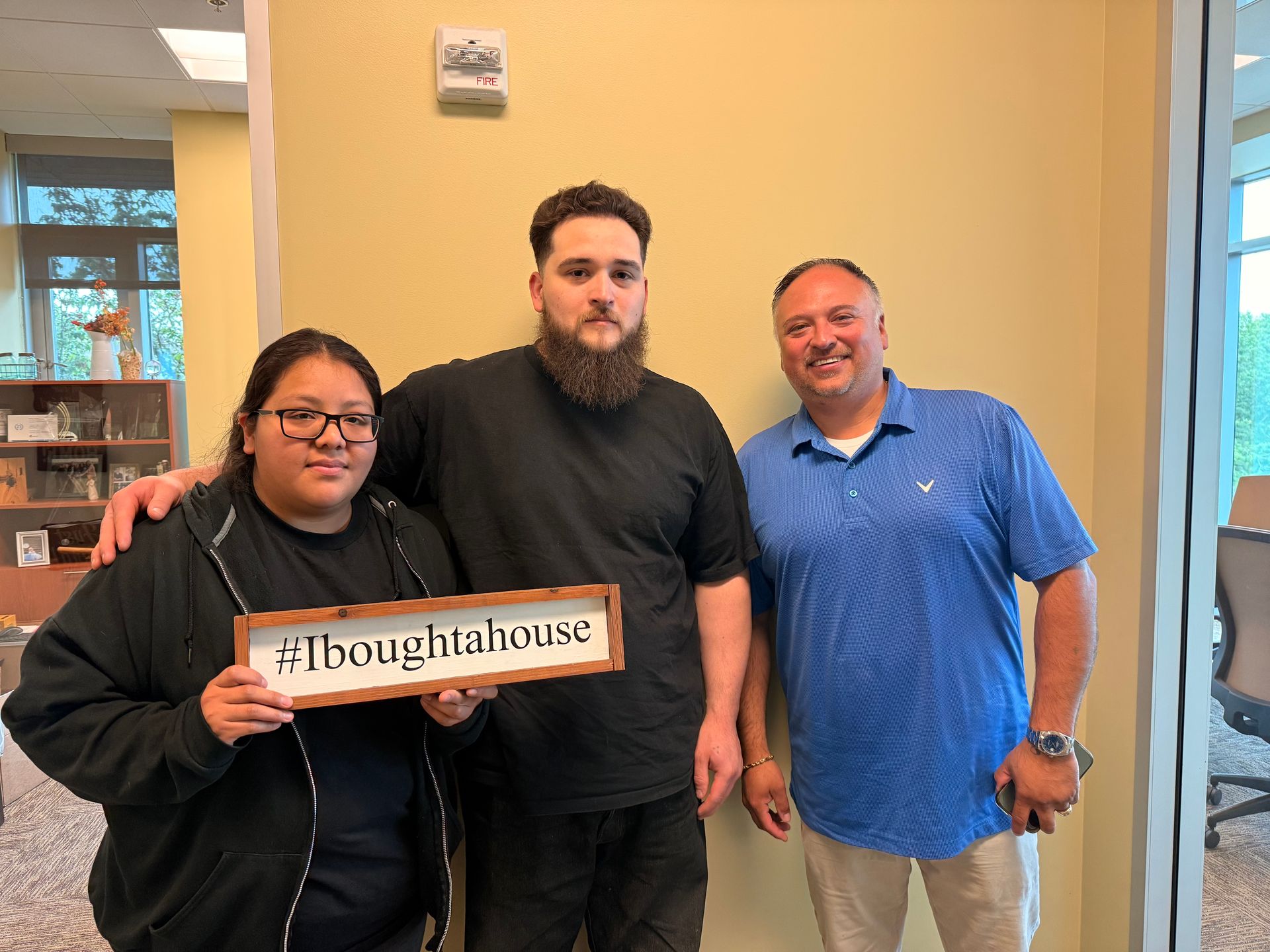 Three people posing: woman holding a sign, two men. Yellow wall, modern office setting.
