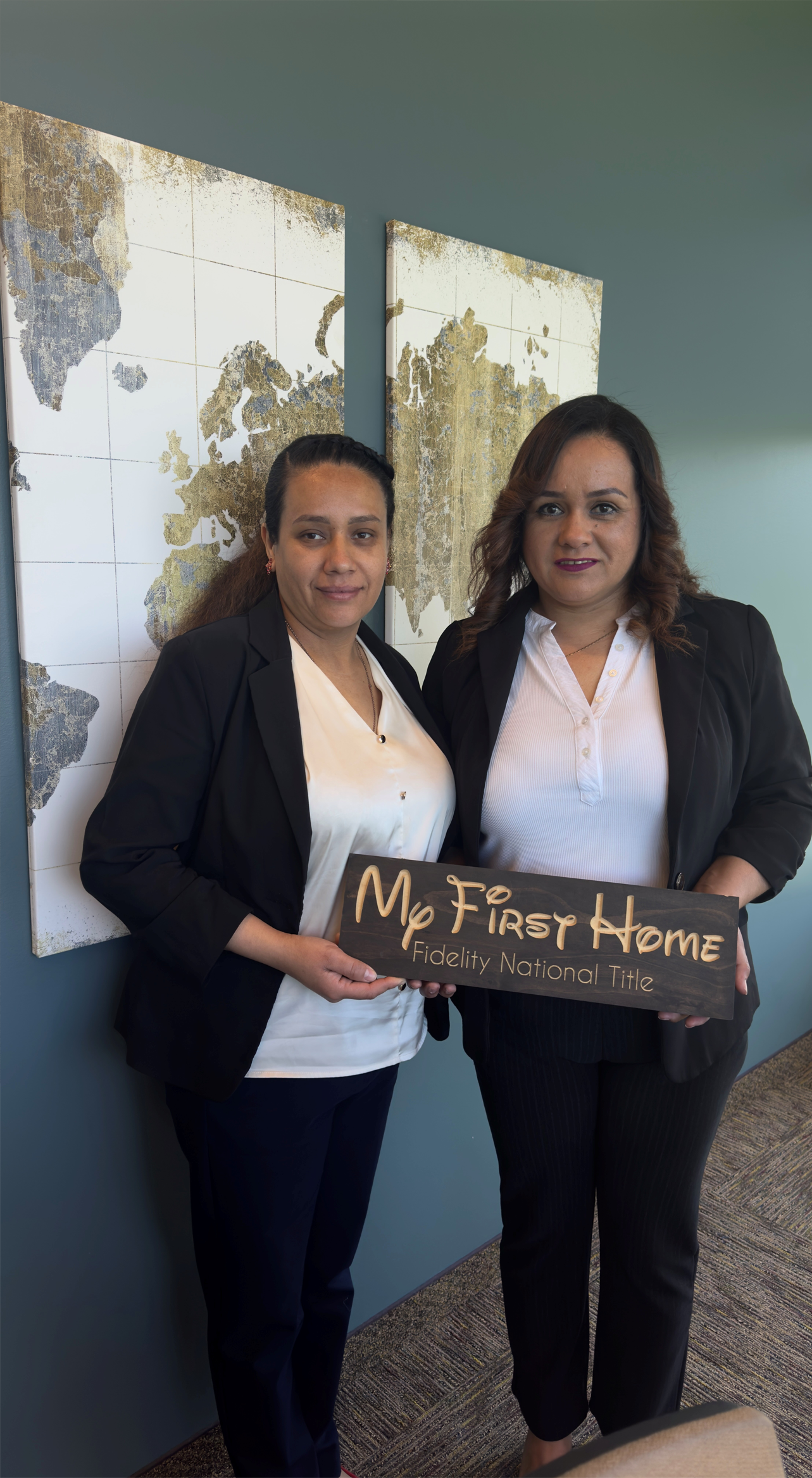 Two women smiling, holding a My First Home sign. Standing near abstract art on a wall.