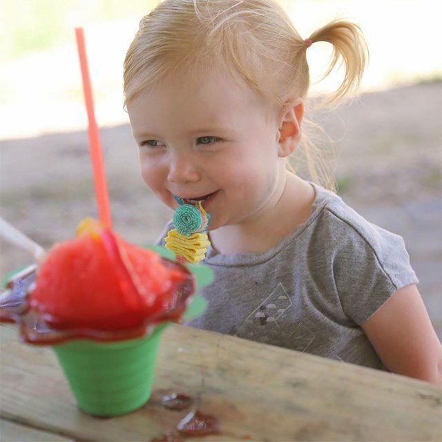 A little girl is sitting at a table with a cup of watermelon shaved ice and a lollipop in her mouth