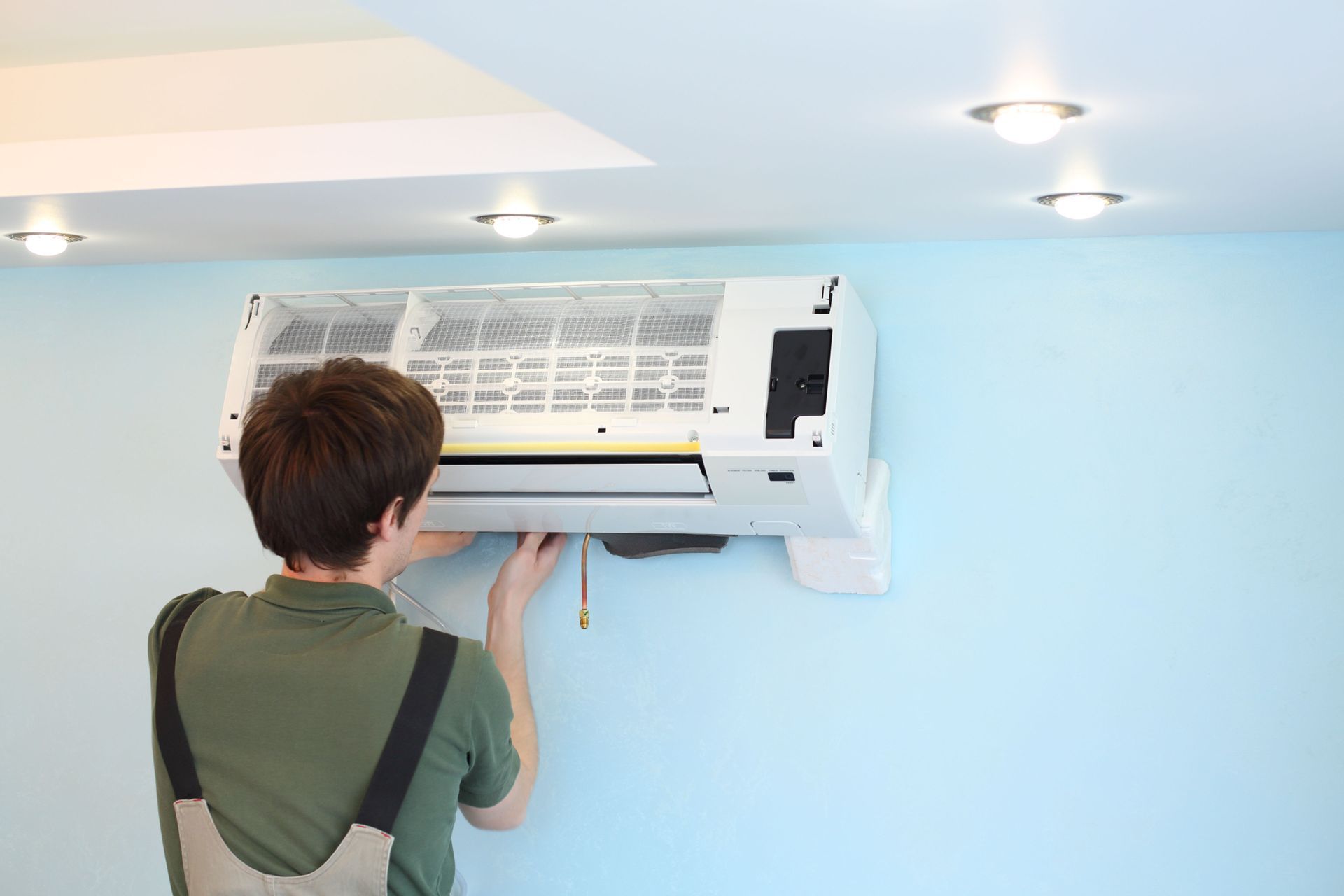 A technician in work coveralls repairs a wall-mounted air conditioning unit in a room with light blue walls.
