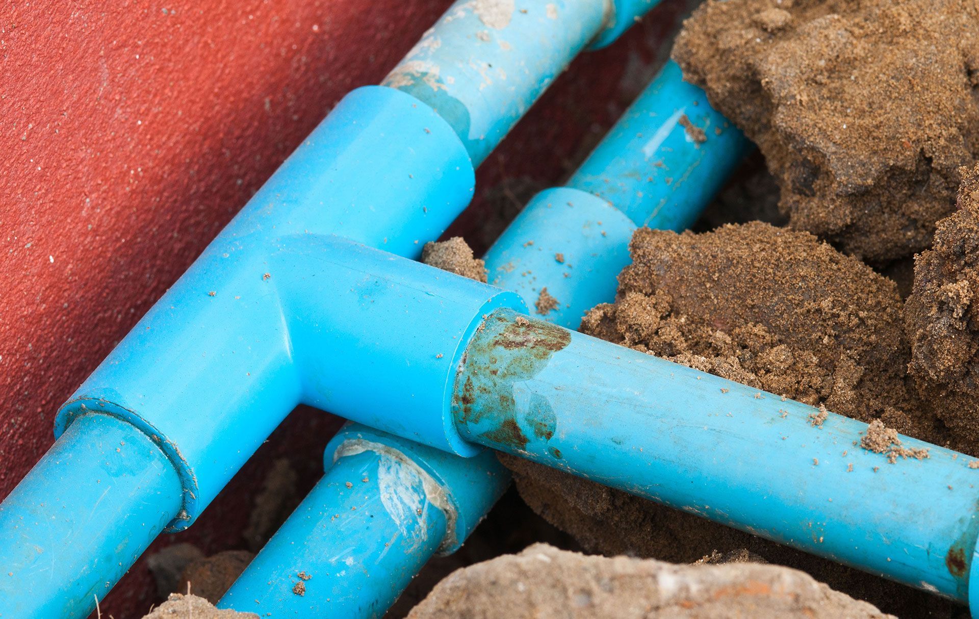 Close-up of blue PVC pipes connected by a T-joint, partially buried in soil next to a red wall.