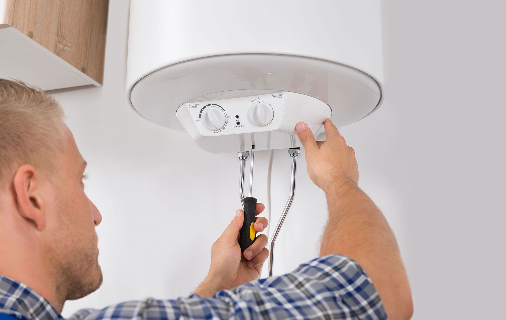 A person in a plaid shirt uses a screwdriver to repair a white wall-mounted electric water heater.