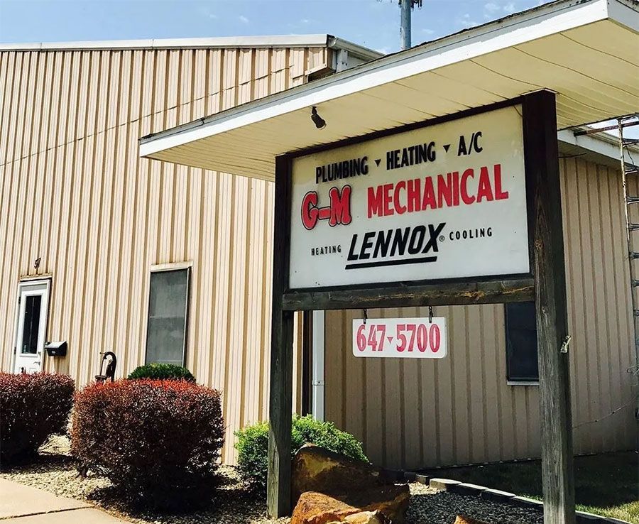 A business sign for C-M Mechanical, specializing in plumbing, heating, and A/C, standing outside a tan metal building.