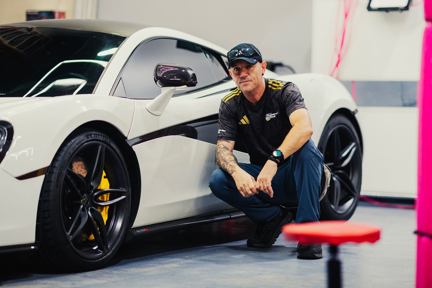 Man crouches beside a white sports car with black accents and tinted windows, yellow brake calipers, in a garage.