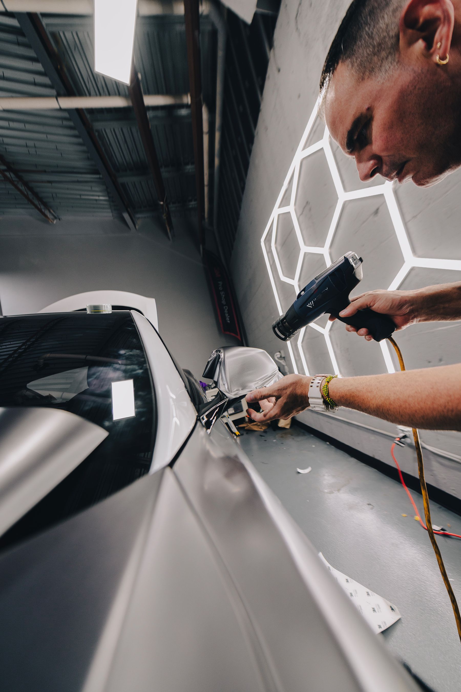 Person applying film to a car's mirror in a well-lit shop, using a heat gun.