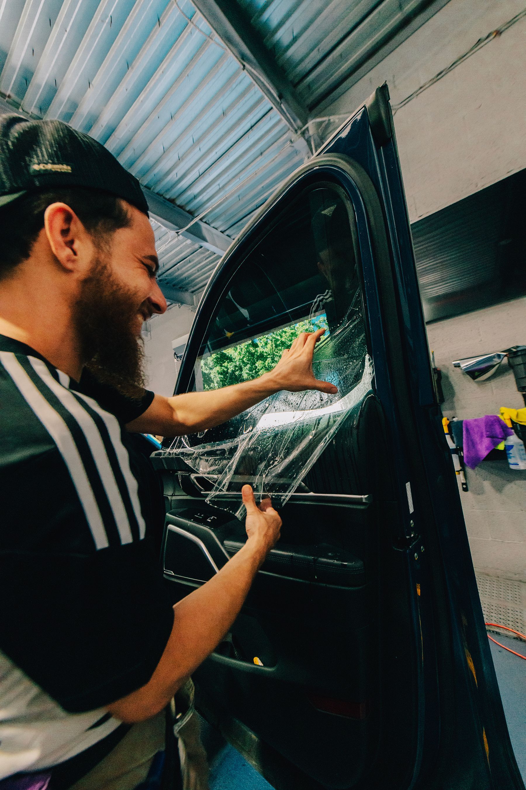 Man applying window tint to a car door, smiling. Workshop setting with visible trees outside.