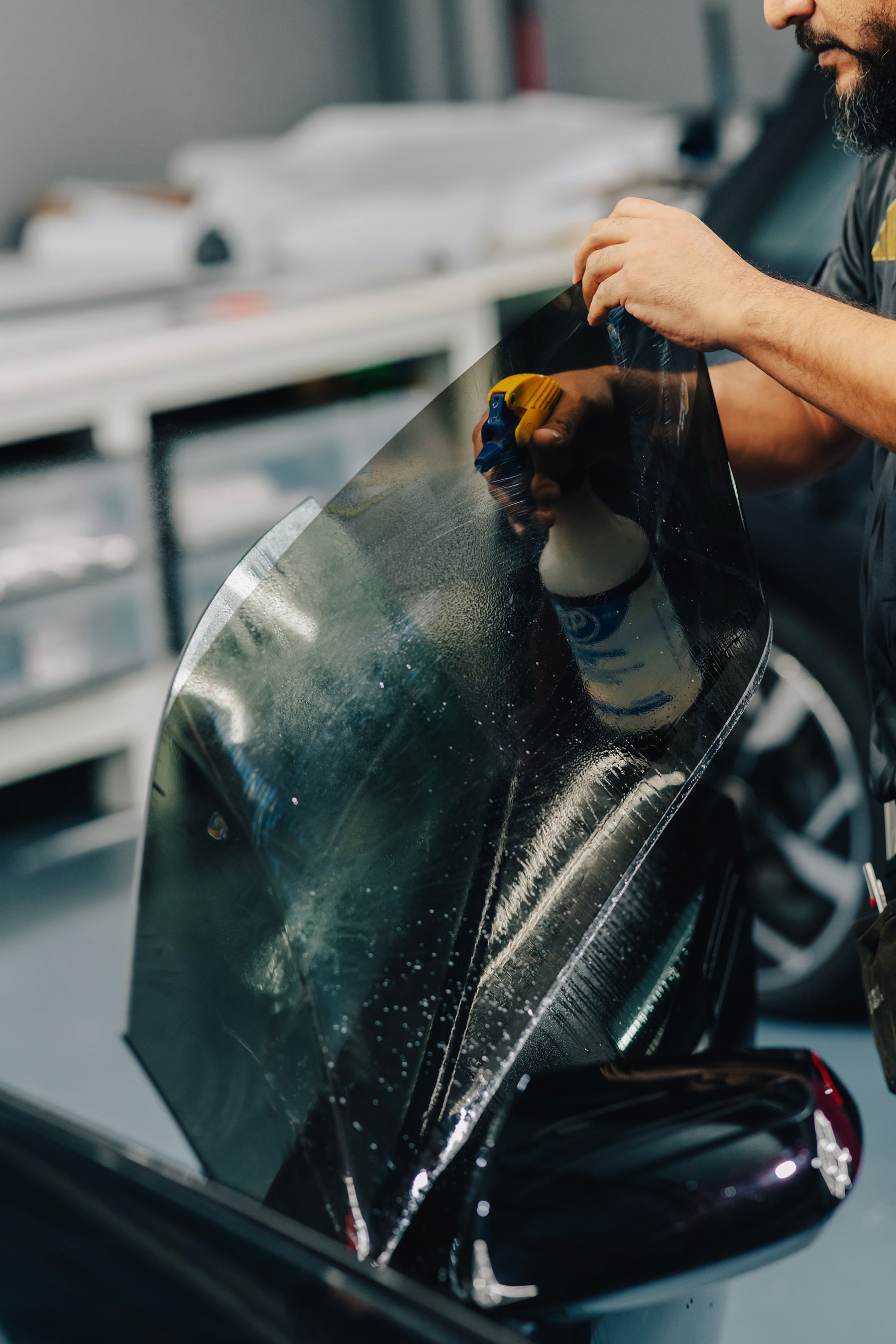 Person spraying tinted film, preparing to apply it to a car window. Workshop setting.