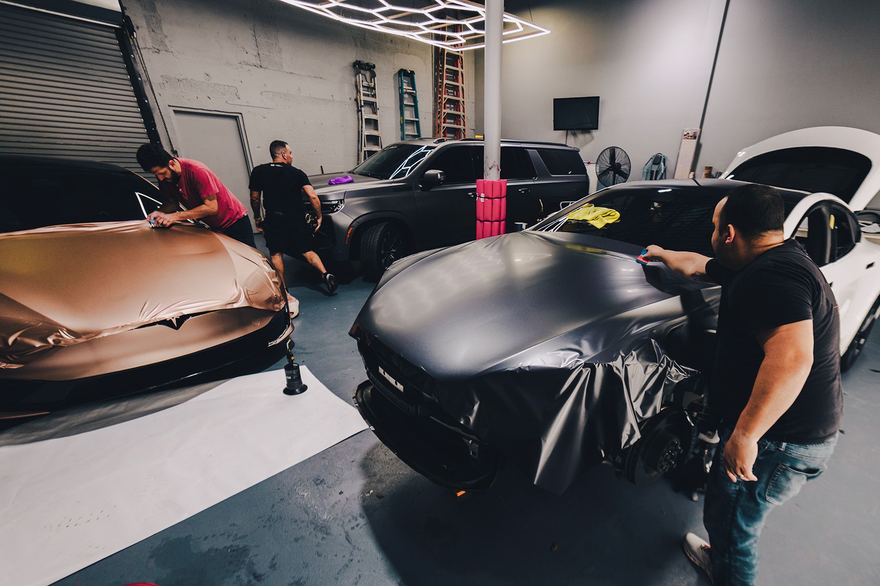 People wrapping cars in a garage. One car has gold wrap, another is black, and another is white.