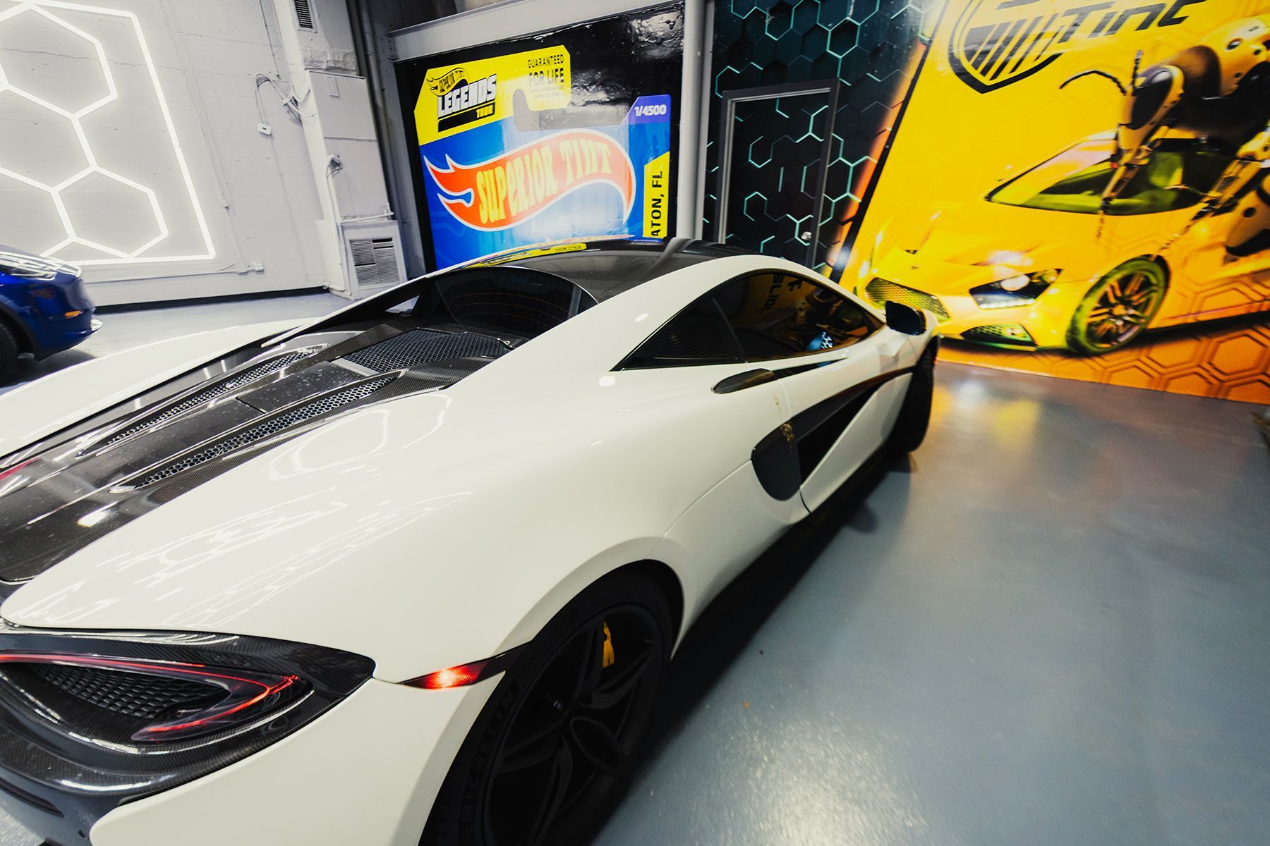 White sports car with black accents in a showroom. Hot Wheels and car tint advertisements in background.