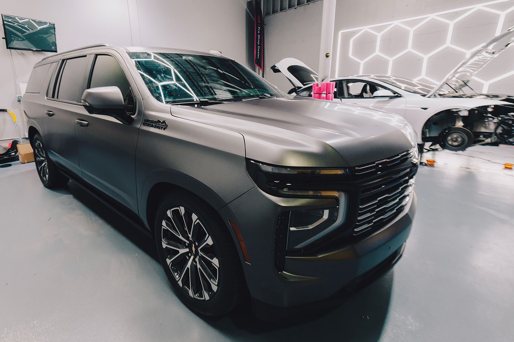 Gray Chevrolet Tahoe in a shop, with a car being worked on in the background.