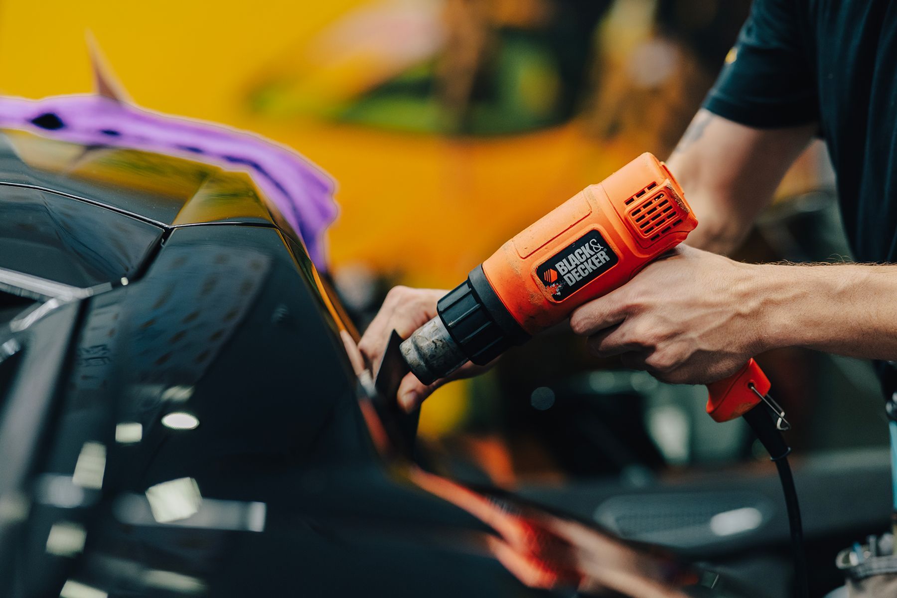 Person using a Black & Decker heat gun to apply film to a black car. Orange tool, indoors.