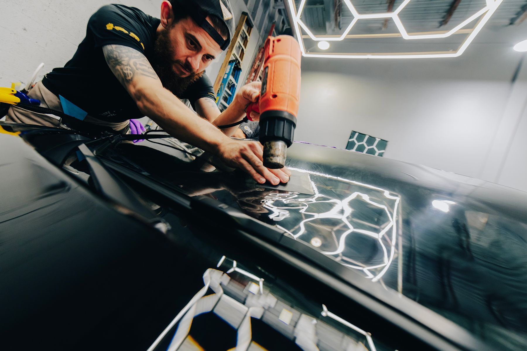 Man applying a wrap to a car roof with a heat gun. Hexagon-shaped lights illuminate the garage.