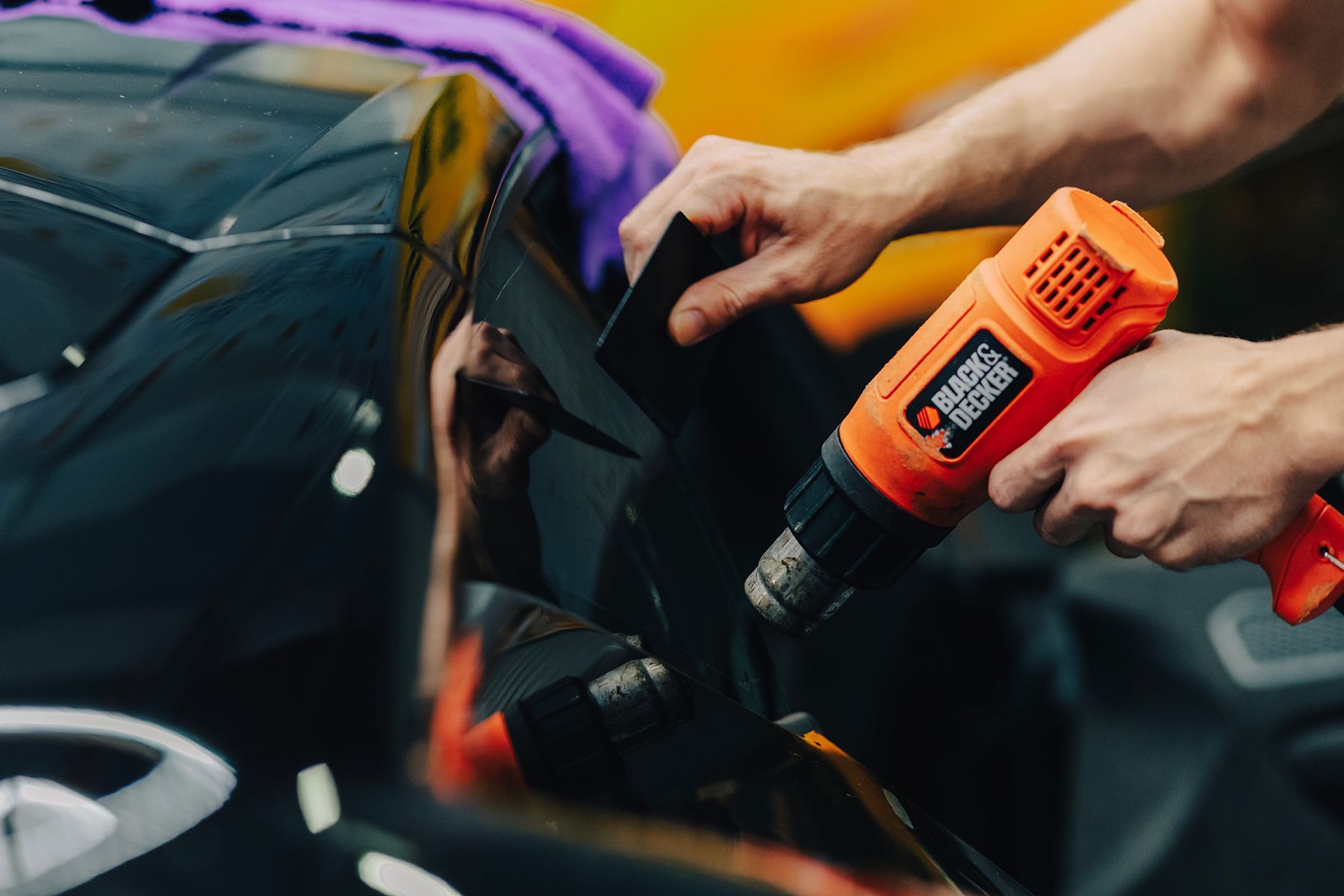 Person uses heat gun and squeegee to apply film to a black car surface.
