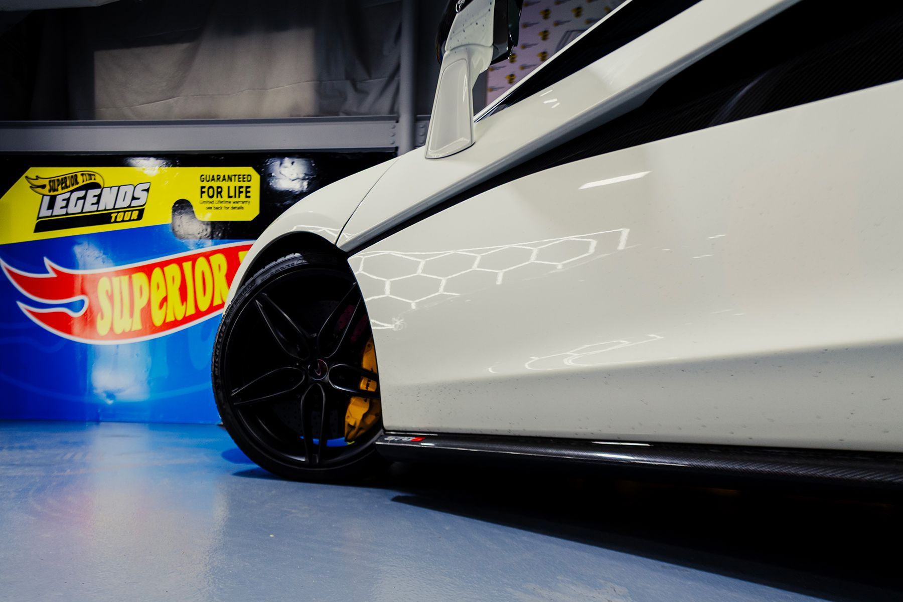 White car with black wheels, yellow calipers, and Hot Wheels Superior Legends logo in a garage.