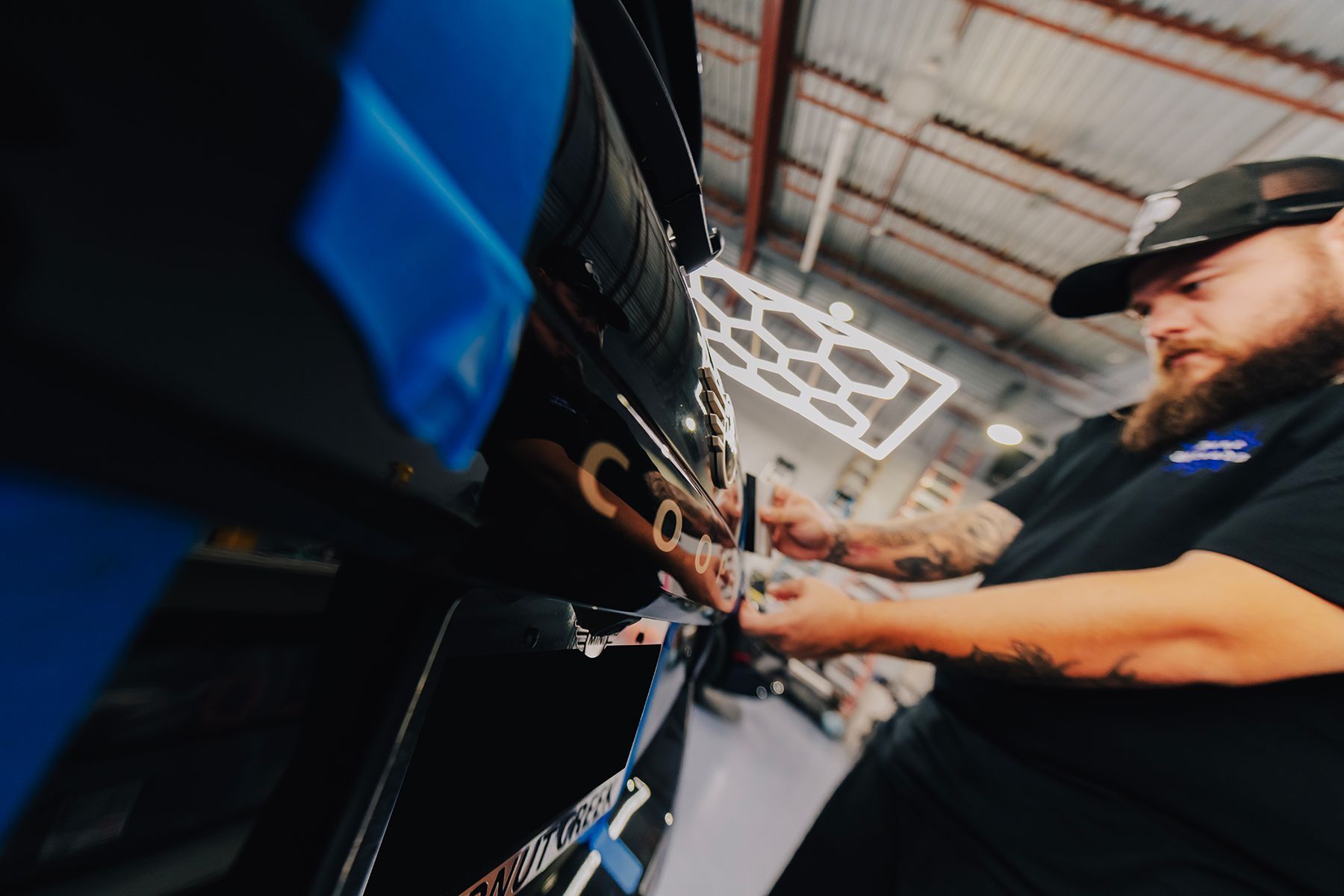 A man installing car wrap in a shop. He uses tools near a black car, wearing a black cap and shirt.