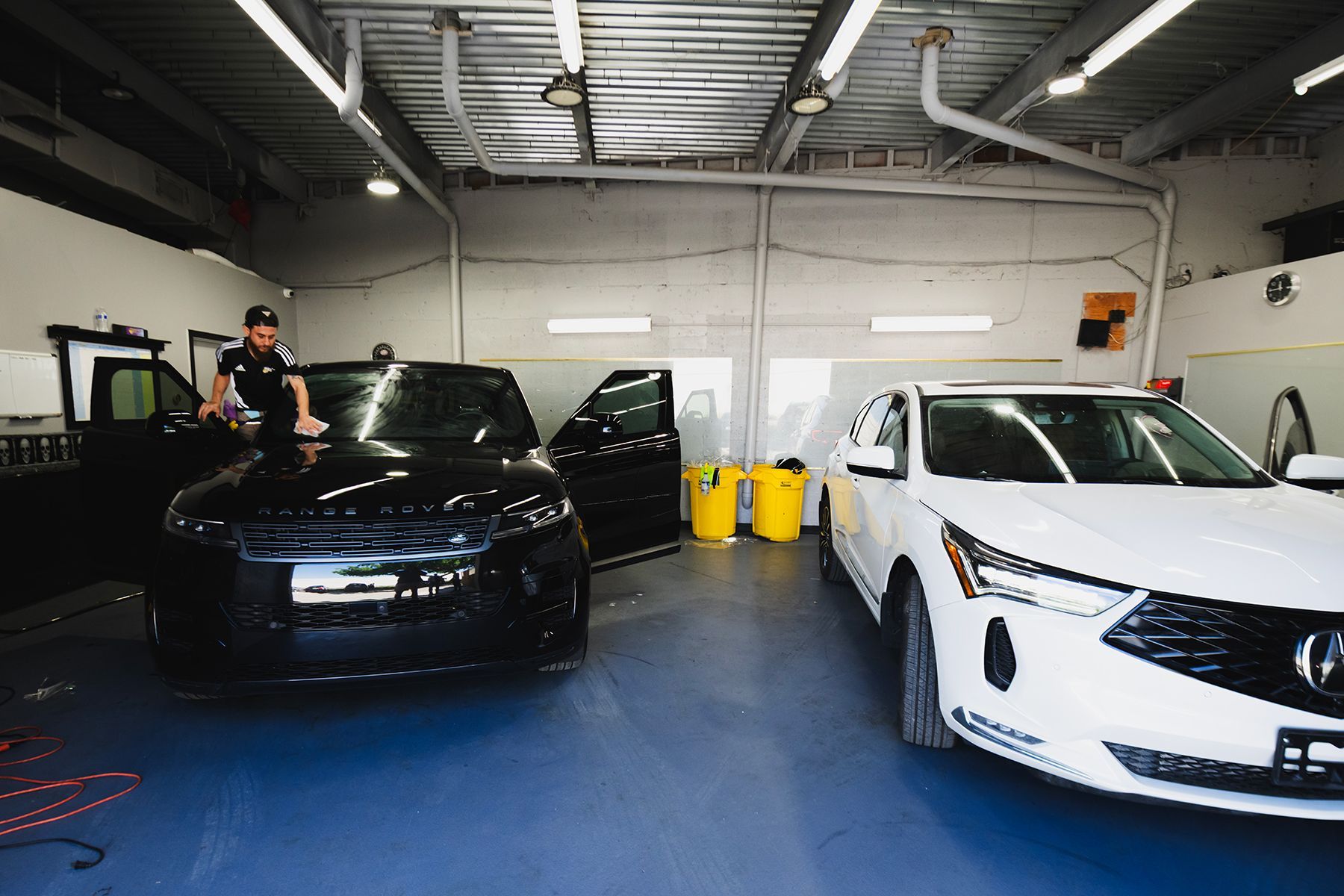 A man cleans a black car in a garage next to a white car.