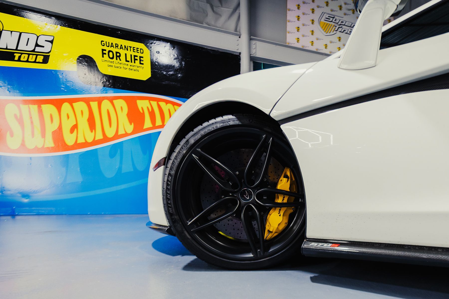 White sports car with black rims and yellow brake calipers; blue backdrop.