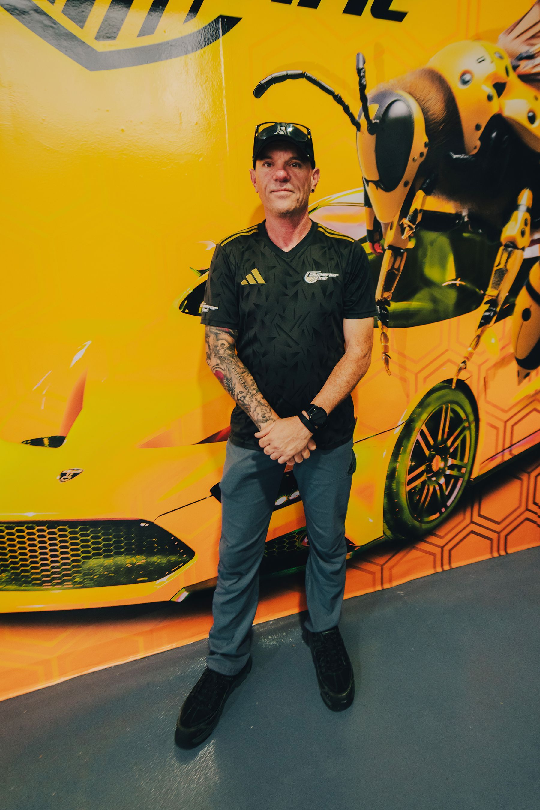 Man with tattoos stands in front of a yellow mural of a car and a bee.