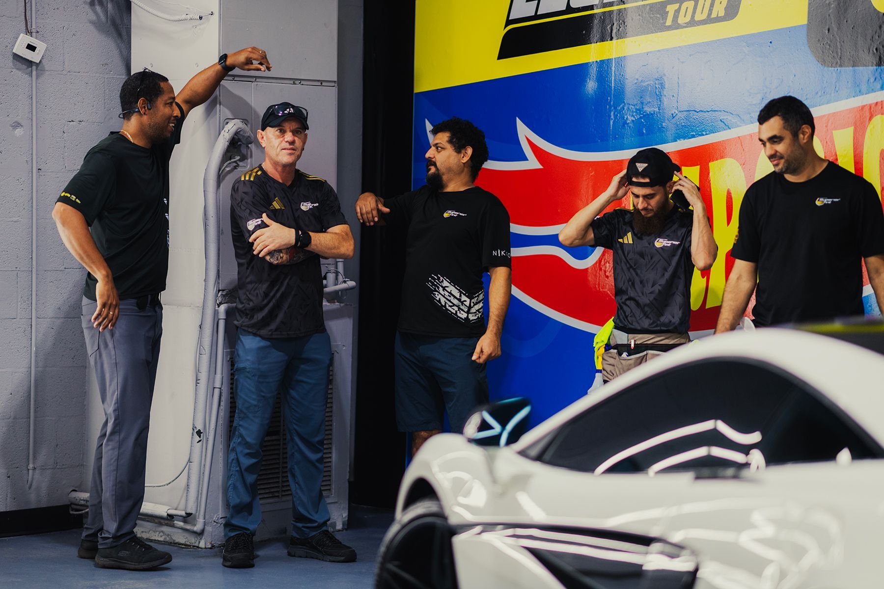 Five men in a garage setting, looking at a white sports car, some gesturing.