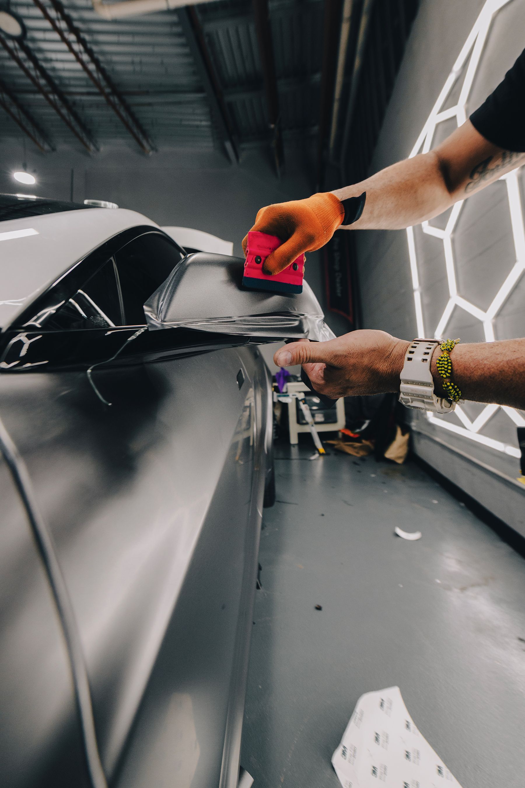 Person wrapping a car in a shop, using a squeegee to apply film. Hands in gloves, grey car, geometric lights.