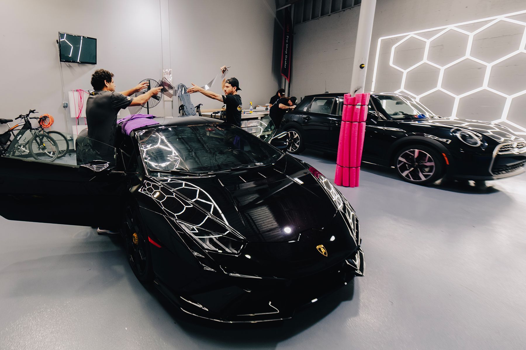 Two people cleaning a black sports car inside a garage; a black Mini Cooper sits nearby.