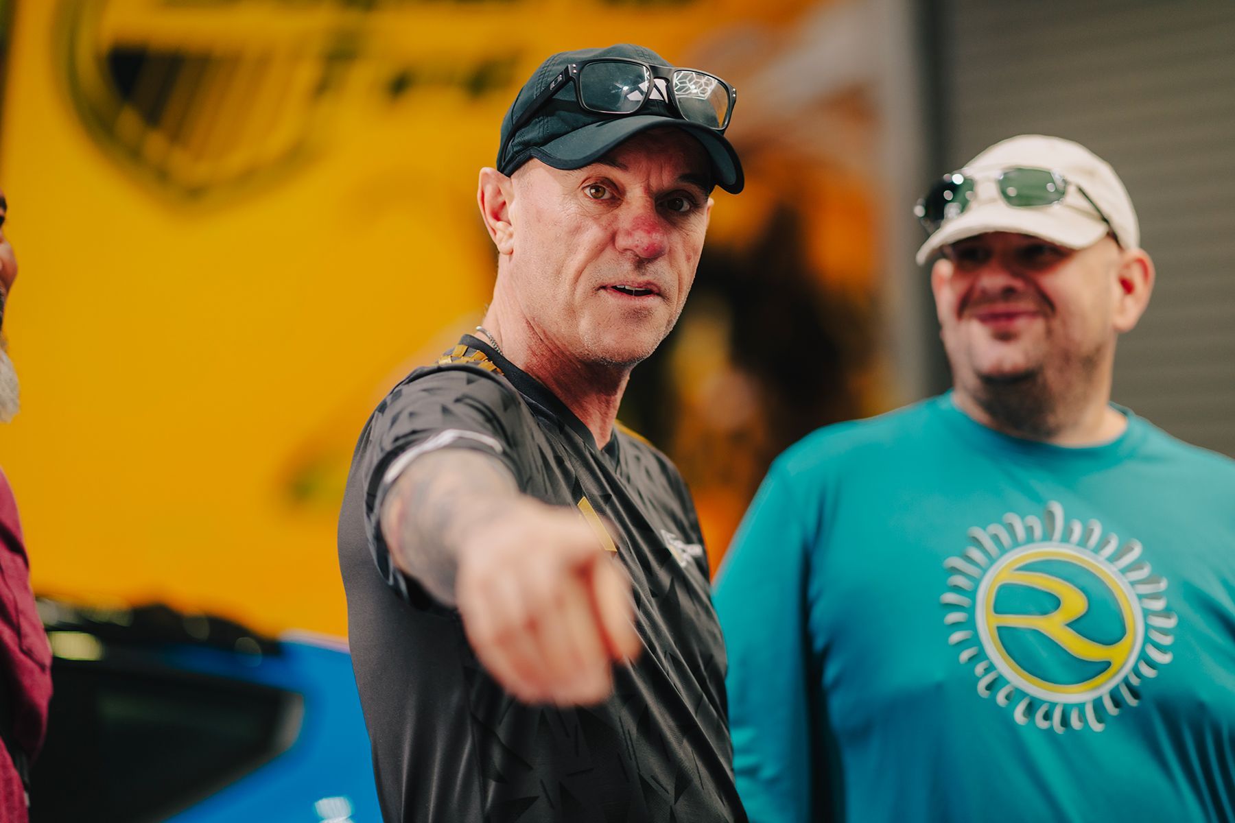 Man in a black cap points toward the left; other men look on. Bright yellow and blue background.