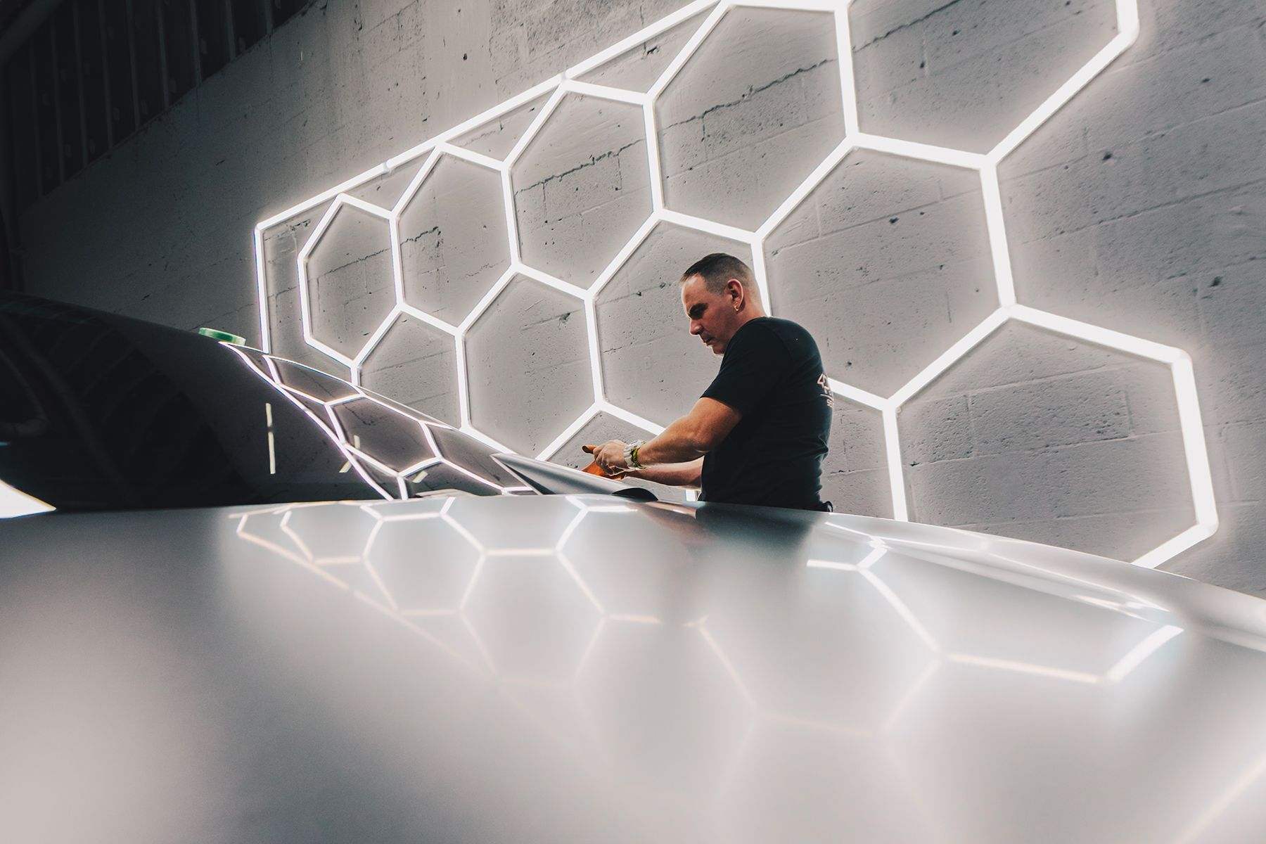 Man writing on a surface, in front of honeycomb-shaped neon lights, with a car in the foreground.