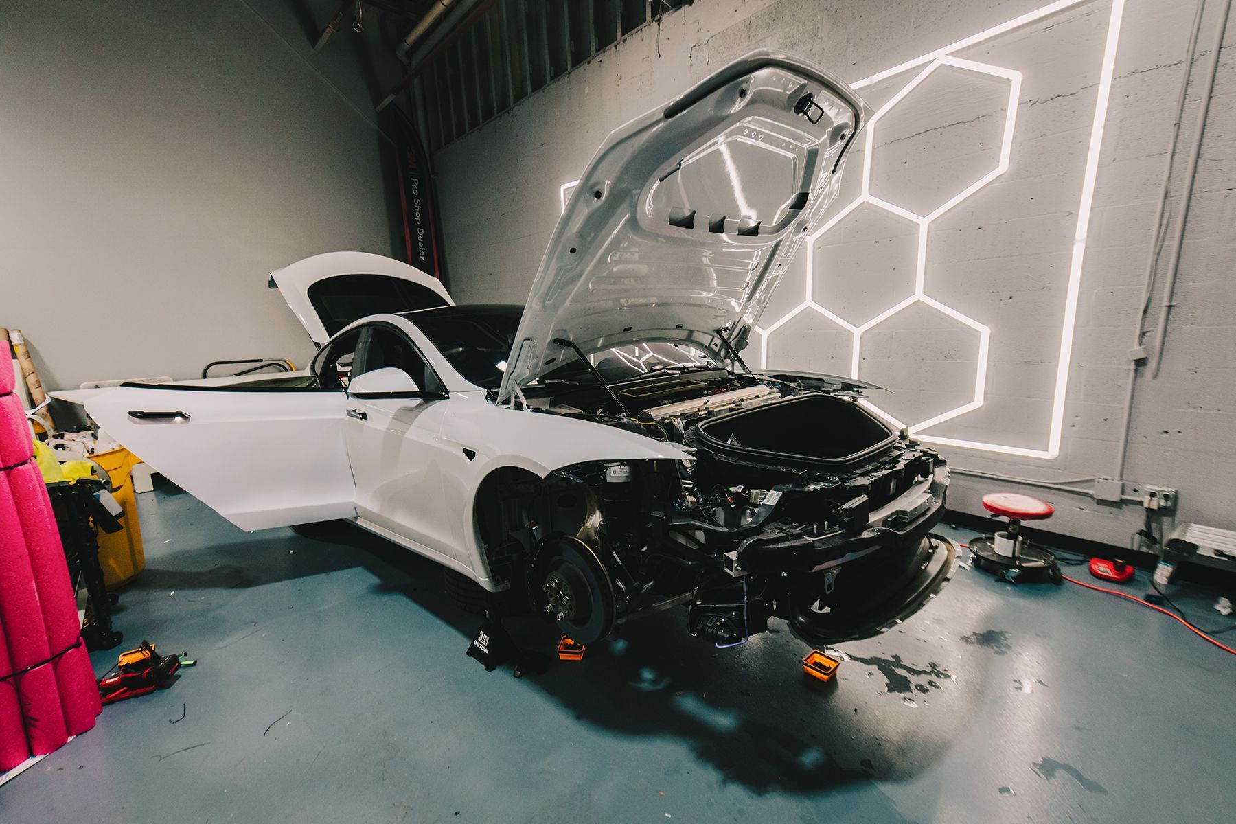 White Tesla car with open doors and hood in a garage; front end is dark, possibly being worked on.