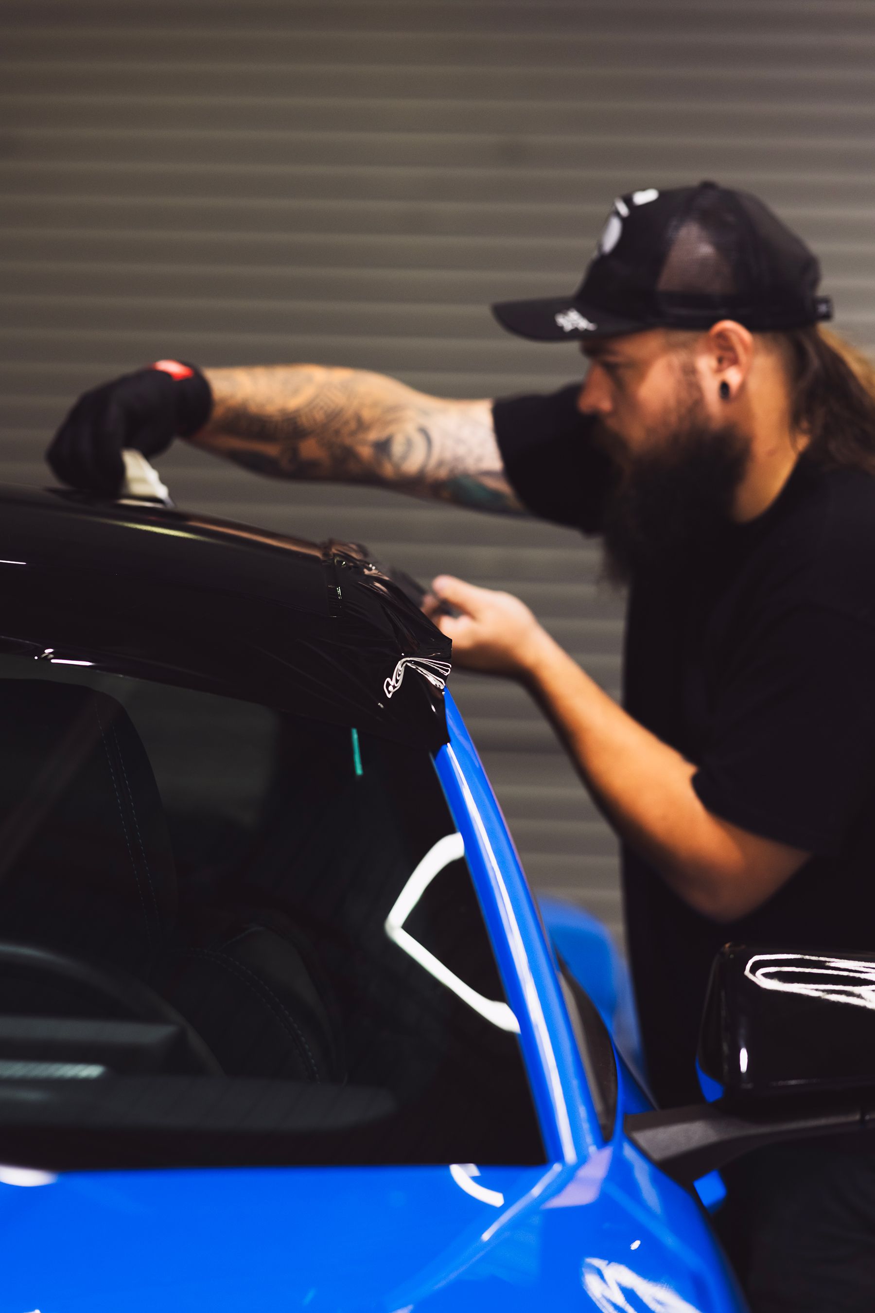 Person applying black vinyl wrap to a blue car's roof in a garage.