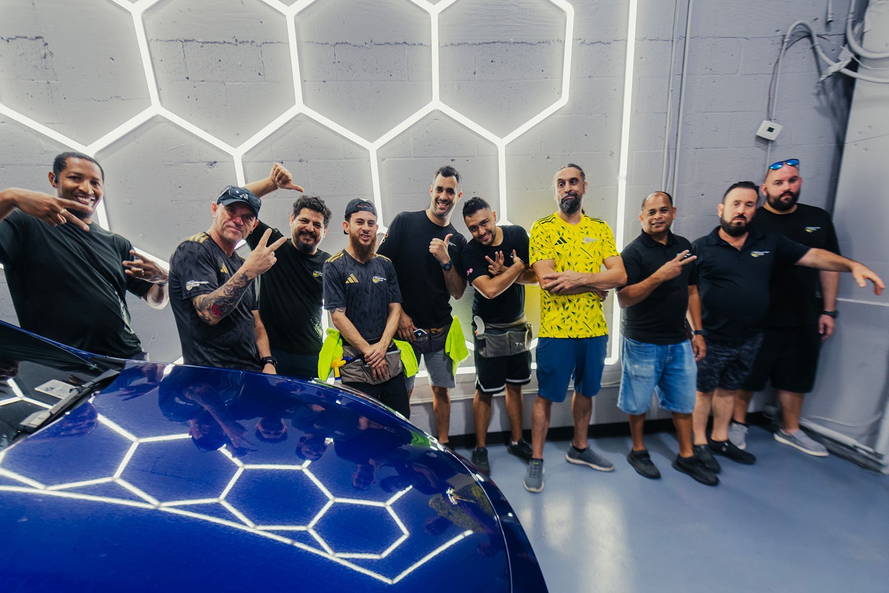 Group of people posing with a blue car in a car detailing shop. Some point to the car; shop has honeycomb lights.