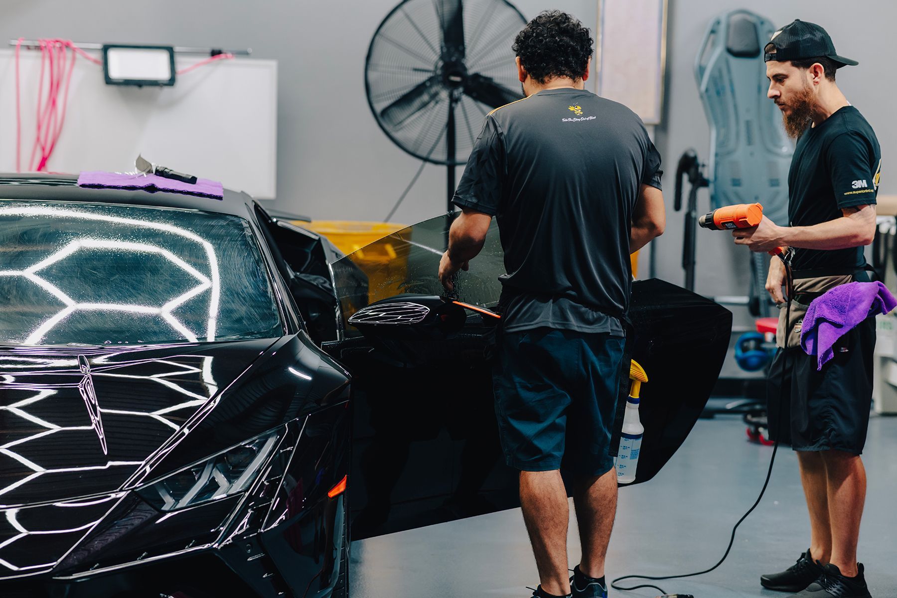 Two people applying tinted film to a black car window in a garage. One holds a heat gun.