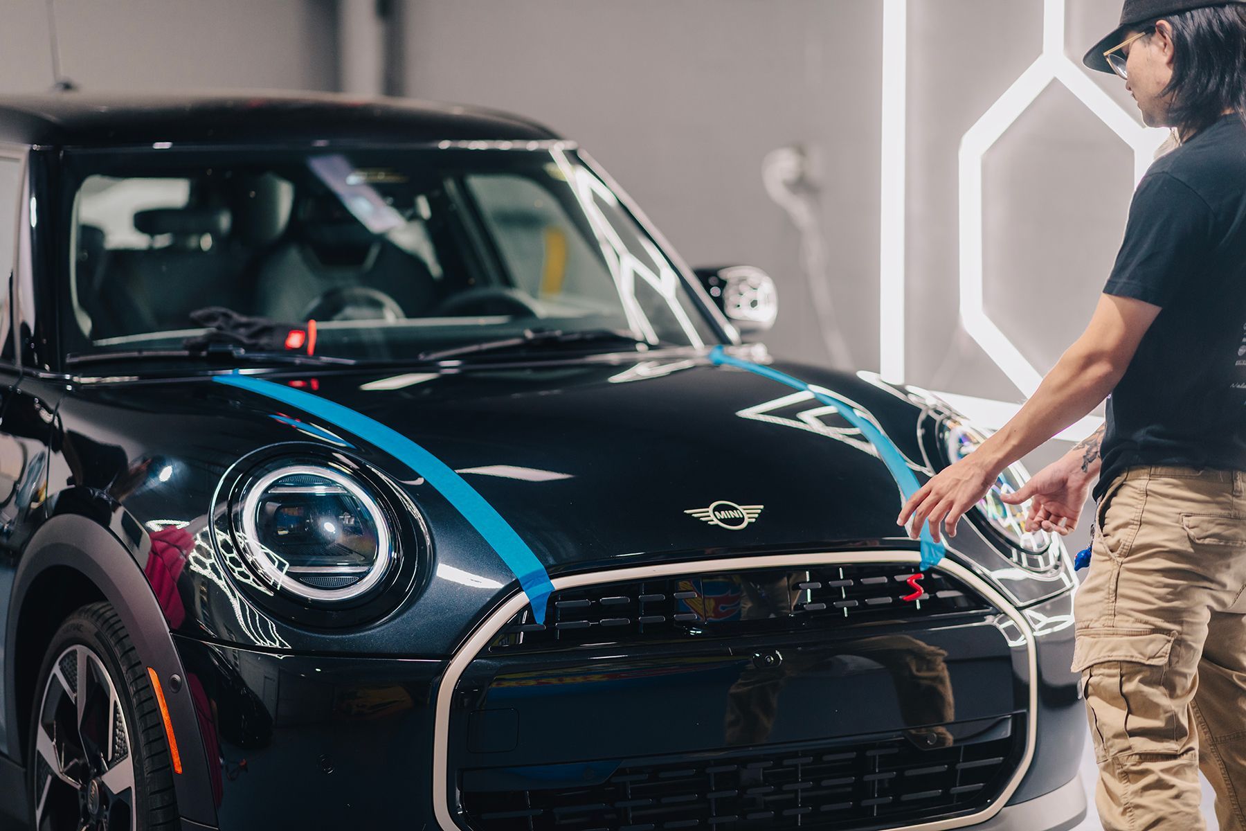 Person in beige pants examines a dark blue MINI Cooper with blue racing stripes in a well-lit garage.