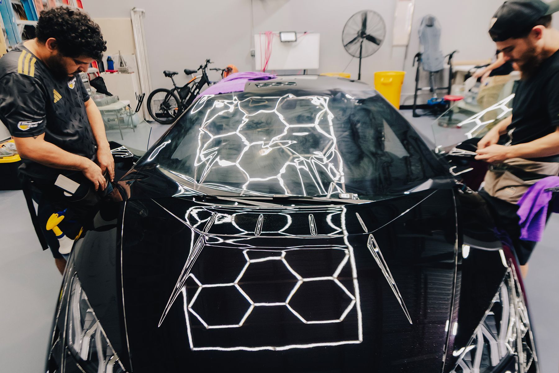 Two people applying film to a black car. They are in a garage-like setting.