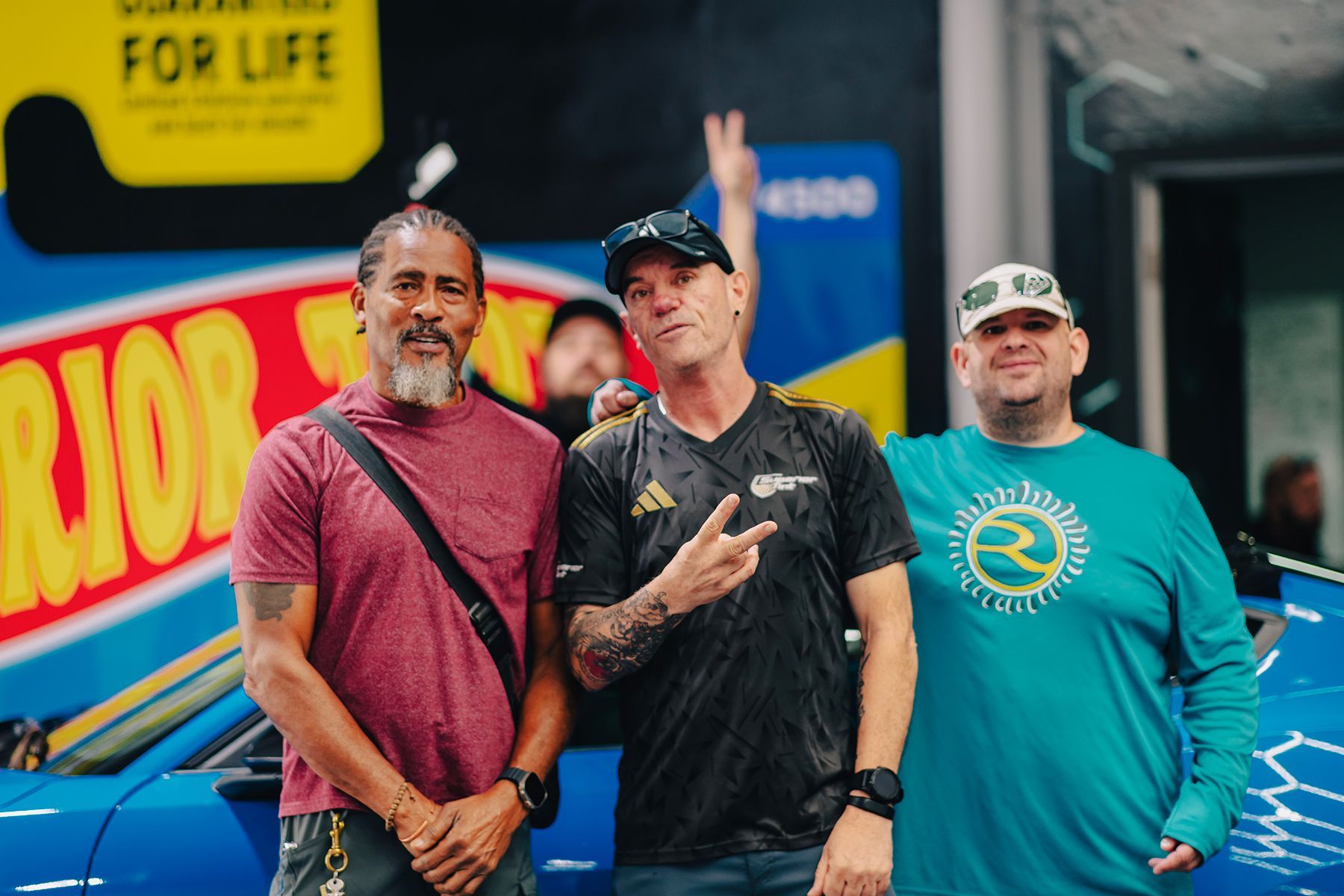 Three men pose in front of a blue car. One man makes a peace sign. They stand near a colorful sign.