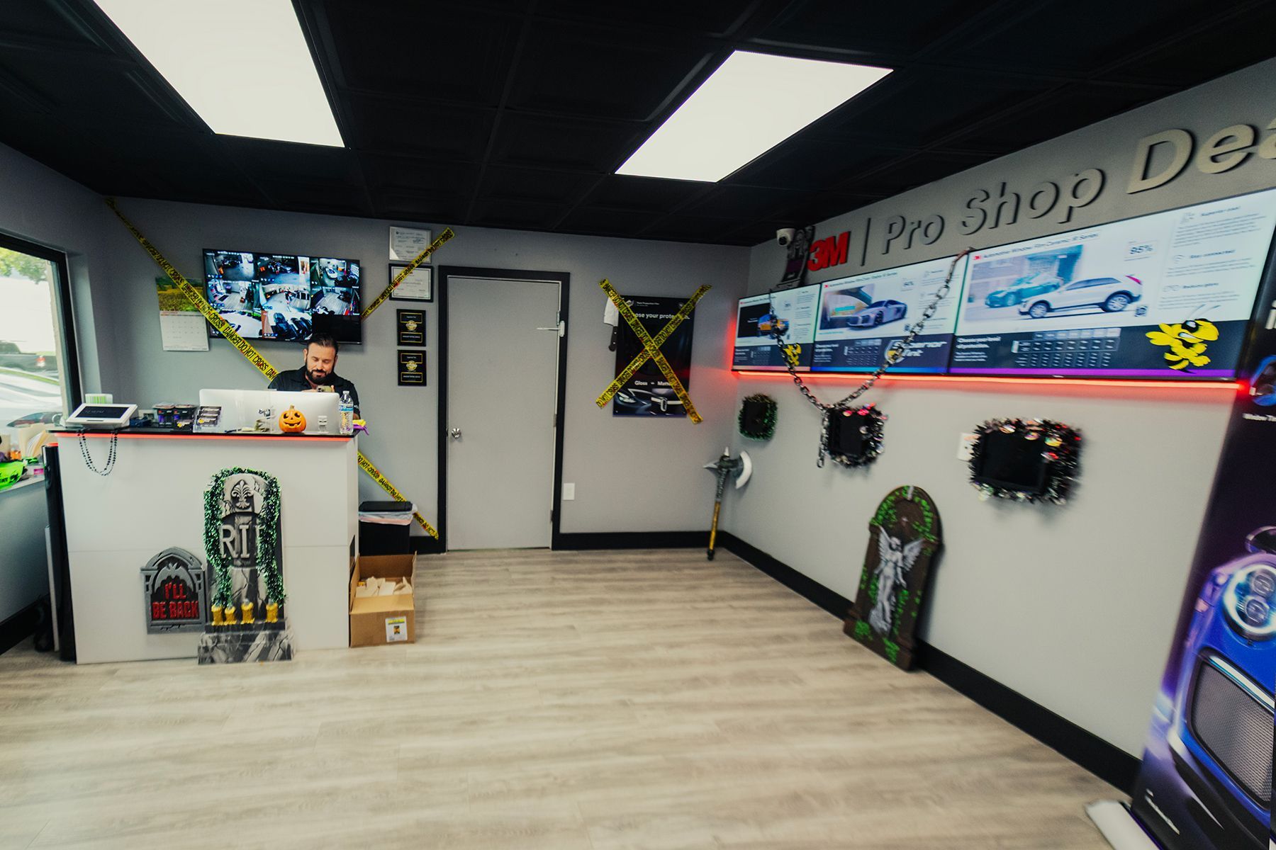 Interior car shop; a man at the front desk. Display screens and themed decorations on the walls. Black ceiling, light wood floor.