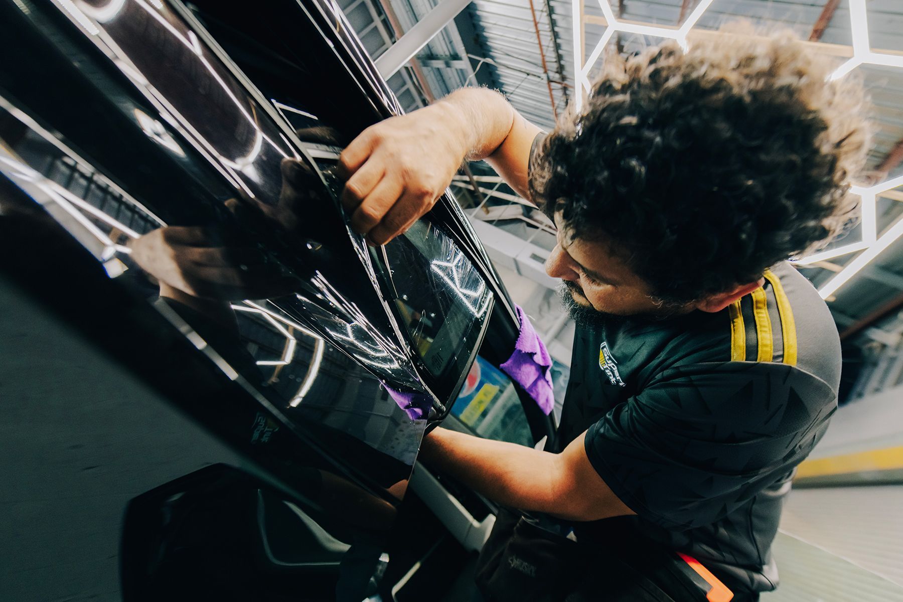 Person applying tint to a black car. They are in a garage and wearing a black shirt with yellow stripes.